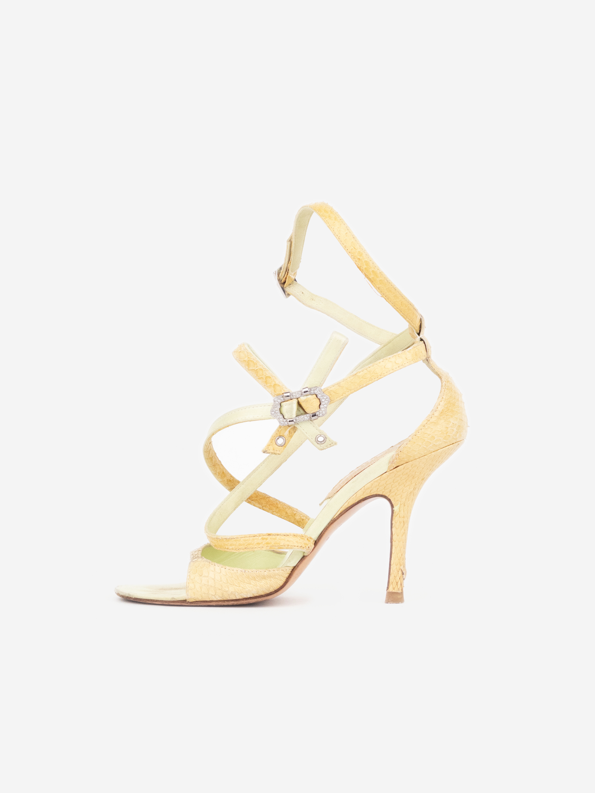 A pale yellow strappy high-heeled sandal with an ankle strap and a small silver-tone heart-shaped charm, shown from the side on a light gray background.